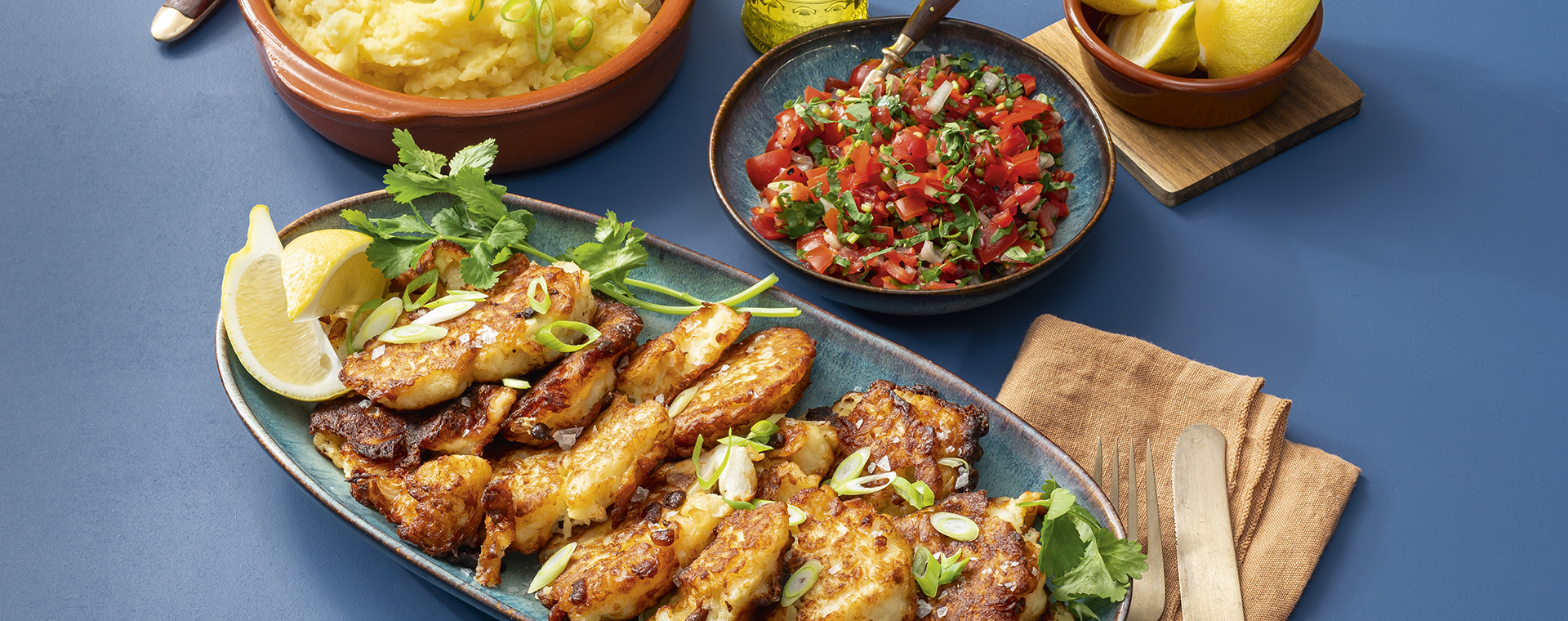 Blumenkohlkrapfen mit frischen Kräutern und Zitronenspalten auf einer Servierplatte, dazu Tomatensalsa und cremiger Kartoffelstampf.