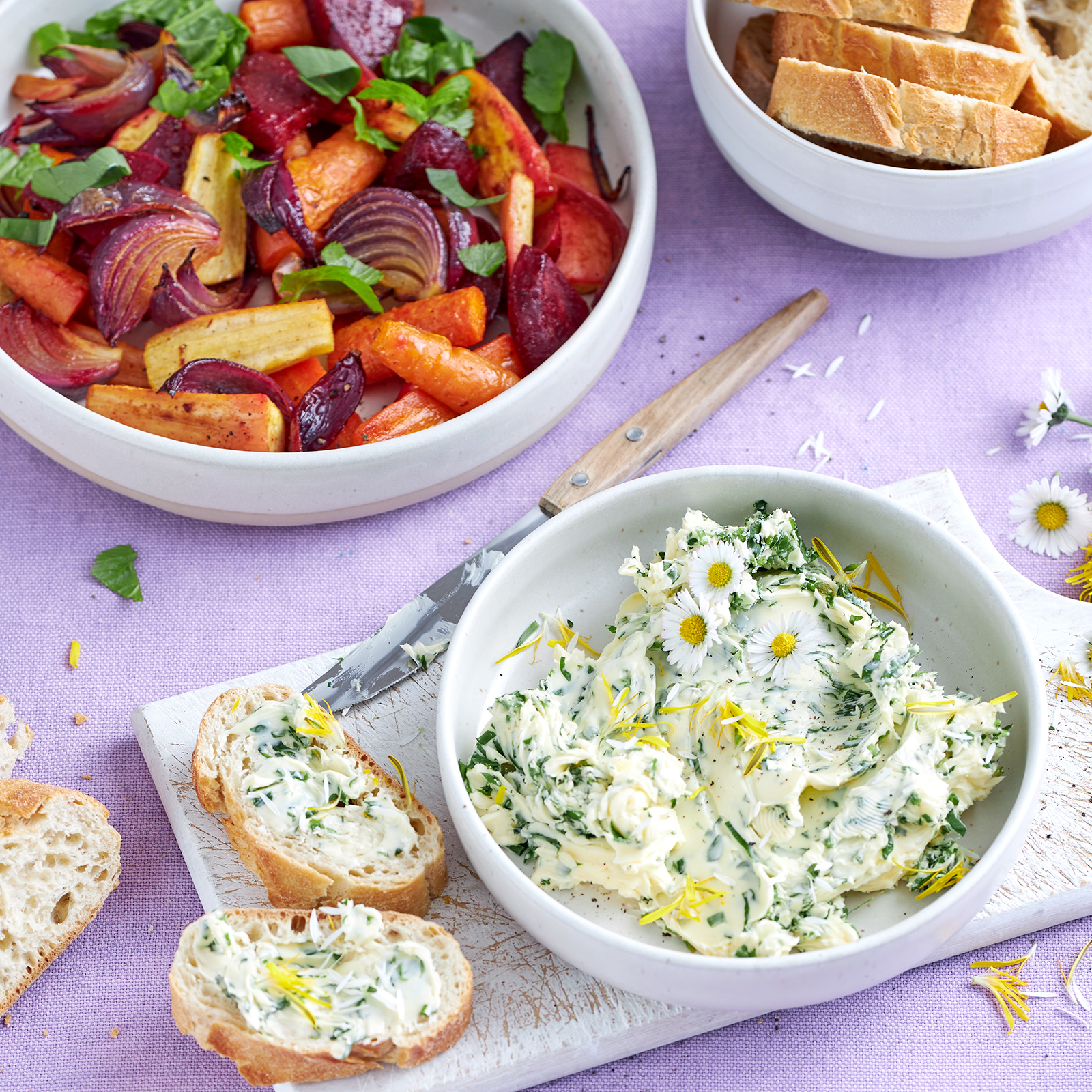 Wildkräuterbutter mit Gänseblümchen in einer Schale, dazu Baguette und Ofengemüse.